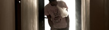A Young Man Of African-American Descent Is Standing In A Hallway, Blowing Vapour Of His E-Cigarette