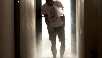 A Young Man Of African-American Descent Is Standing In A Hallway, Blowing Vapour Of His E-Cigarette