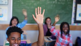Smiling diverse multi-cultural students raising hands in classroom