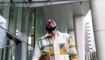 Cheerful man holding smart phone laughing by glass building