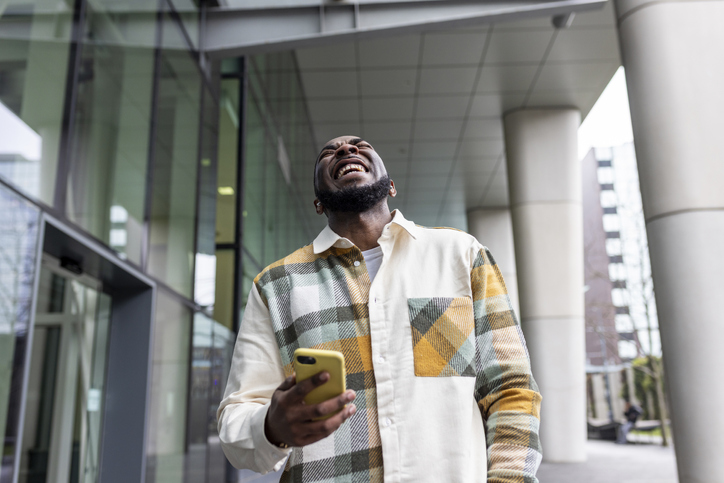Cheerful man holding smart phone laughing by glass building
