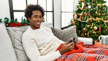 Young african american man playing video game sitting on the sofa by christmas tree at home.