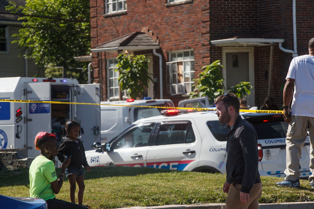 A woman sits with her child behind police tape watching the...