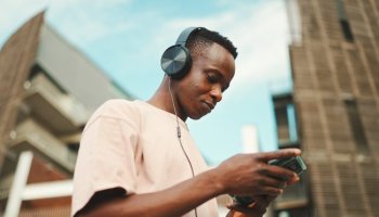 Young african student sitting outside of university wearing headphones, using phone, listening to music