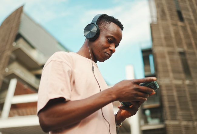 Young african student sitting outside of university wearing headphones, using phone, listening to music