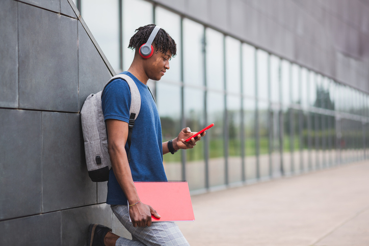 Young man with red headphones and a notebook leaning against the wall, near the entrance door of the university.