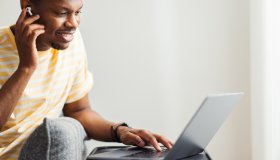 Smiling African American Man Putting Earbuds in His Ear and Using His Laptop Computer at Home