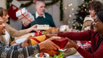 Multi-ethnic family exchanging presents during Christmas party at home. Attractive diverse people receive gift from relatives while having dinner celebrate holiday Thanksgiving on dining table.