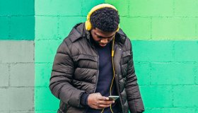 Black African American boy on green background wall. Listening to music with yellow headphones and his mobile phone.