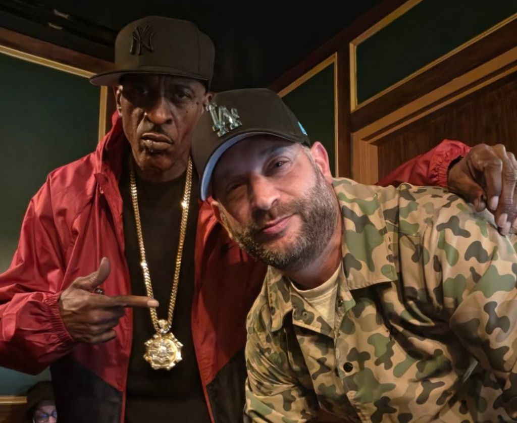 Two men wearing red and camouflage clothing, one with a Yankees cap, posing together in a room with wooden paneling.