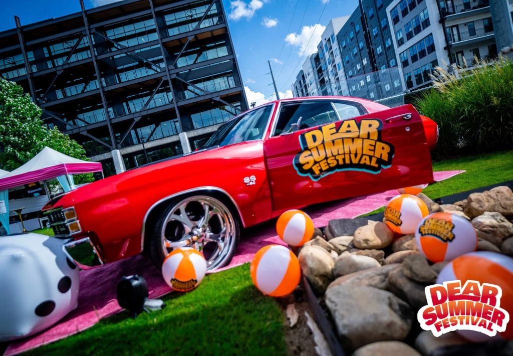 A red car with "Dear Summer Festival" written on the side, surrounded by beach balls and rocks on a grassy field in front of a modern building.