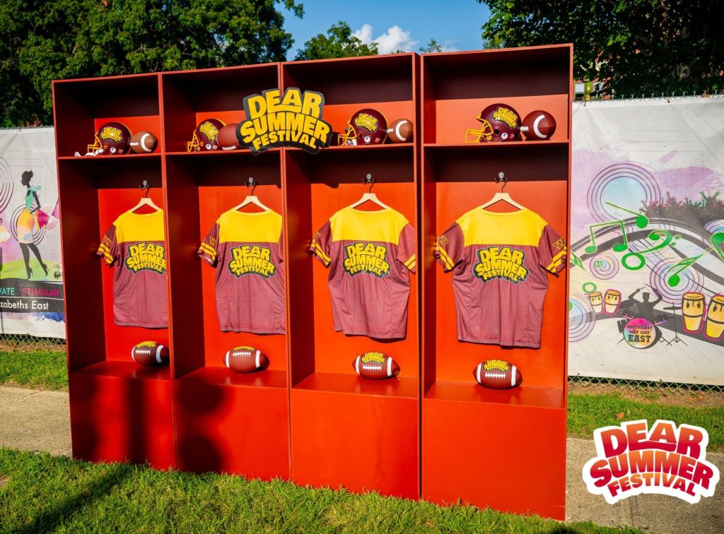 A display of merchandise for the "Dear Summer Festival" event, featuring yellow t-shirts, footballs, and other sports-themed items on red shelves.