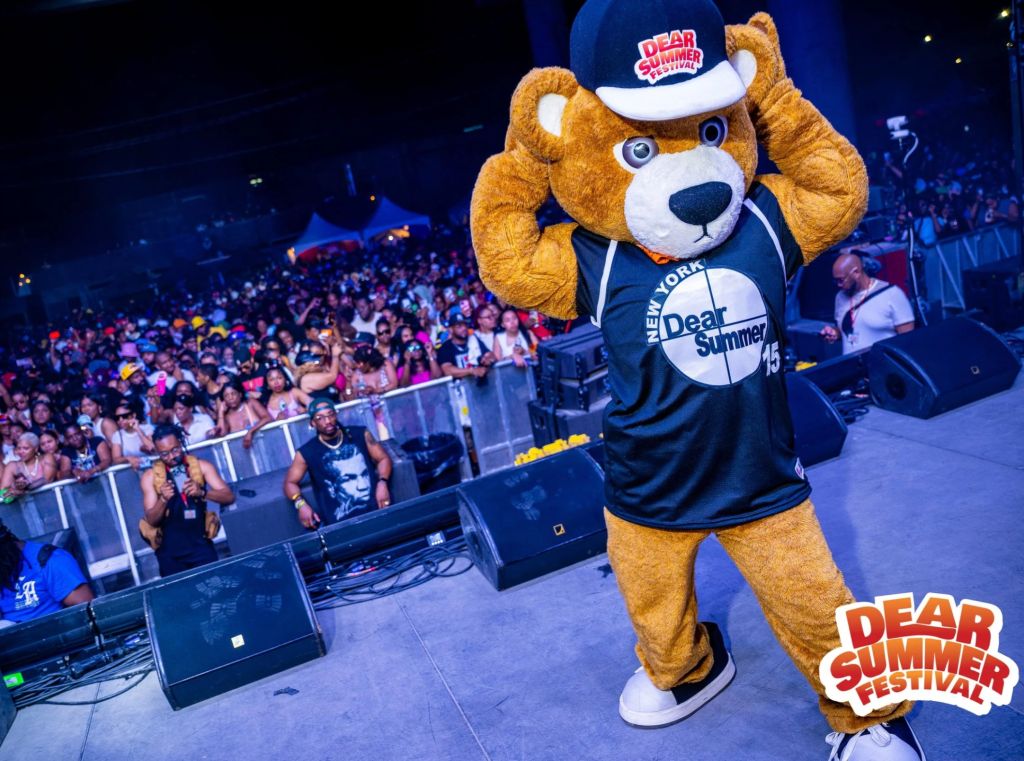 A large crowd at a music festival, with a costumed mascot character wearing a "Dear Summer Festival" jersey and hat standing on stage in front of the audience.