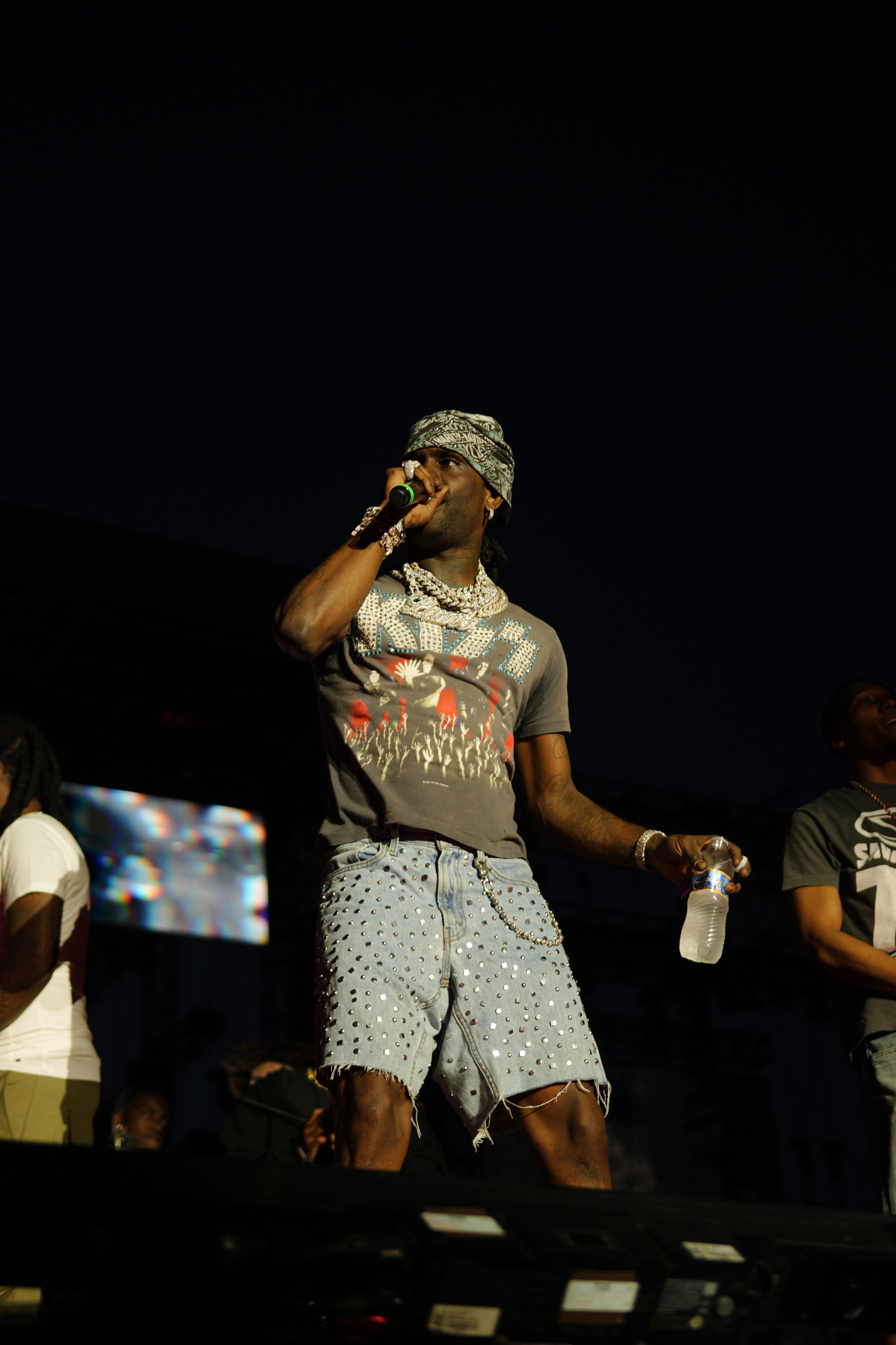 A performer wearing a patterned shirt and hat holding a microphone on stage in front of a dark background.