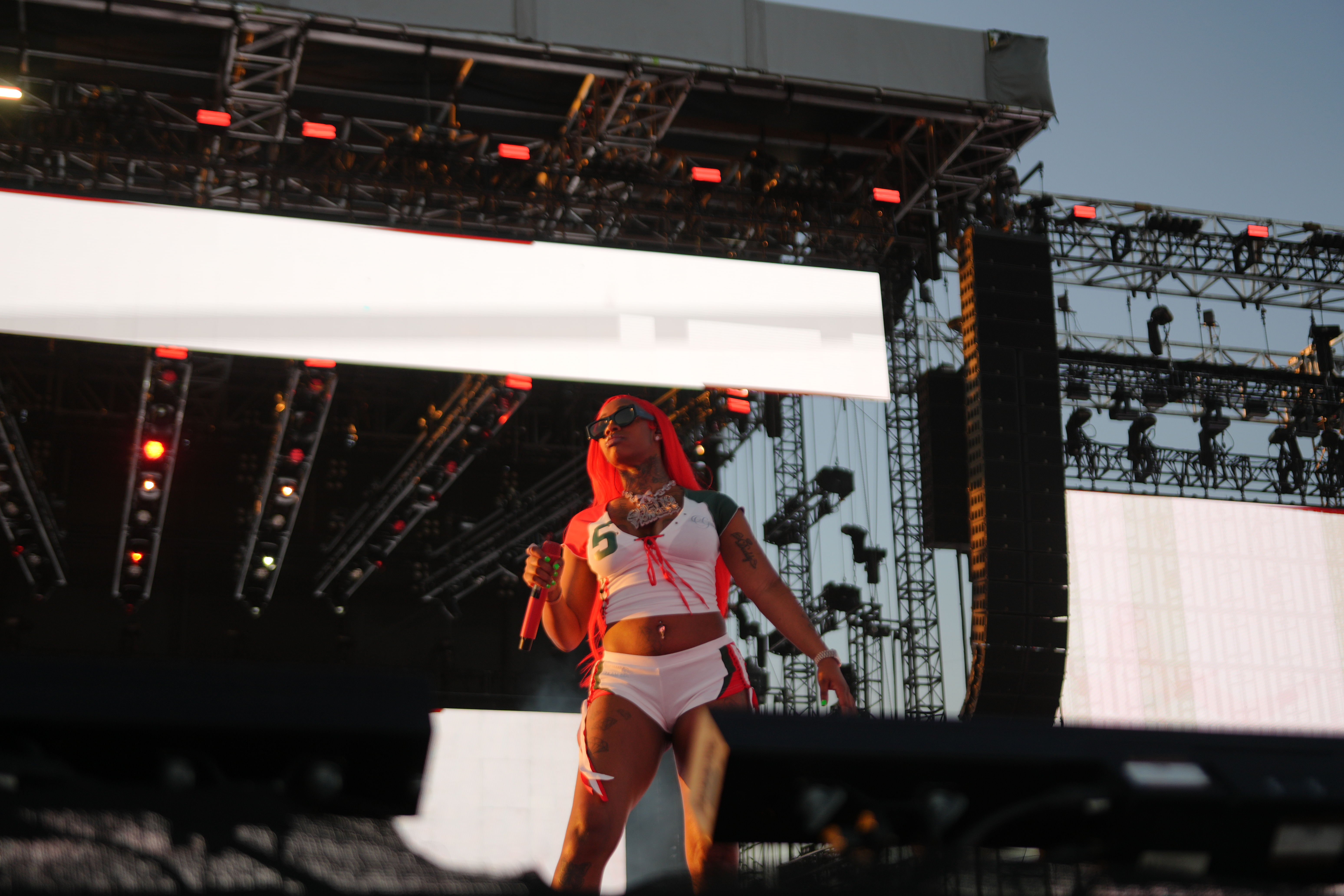 Performer on stage wearing white outfit with red accents, surrounded by lighting and stage equipment.