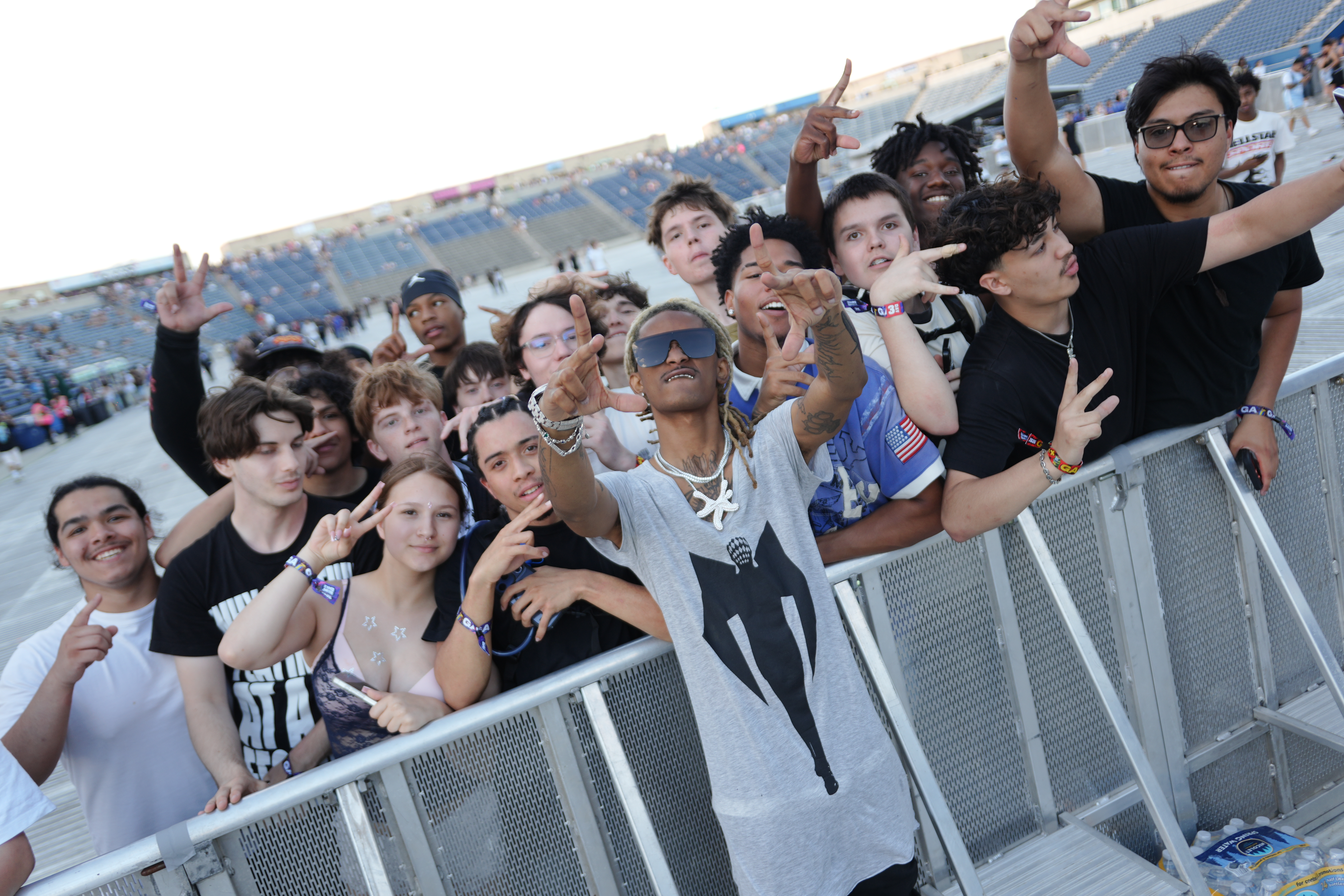 A large crowd of people, mostly young, gathered at an outdoor event or concert, raising their hands and making peace signs.