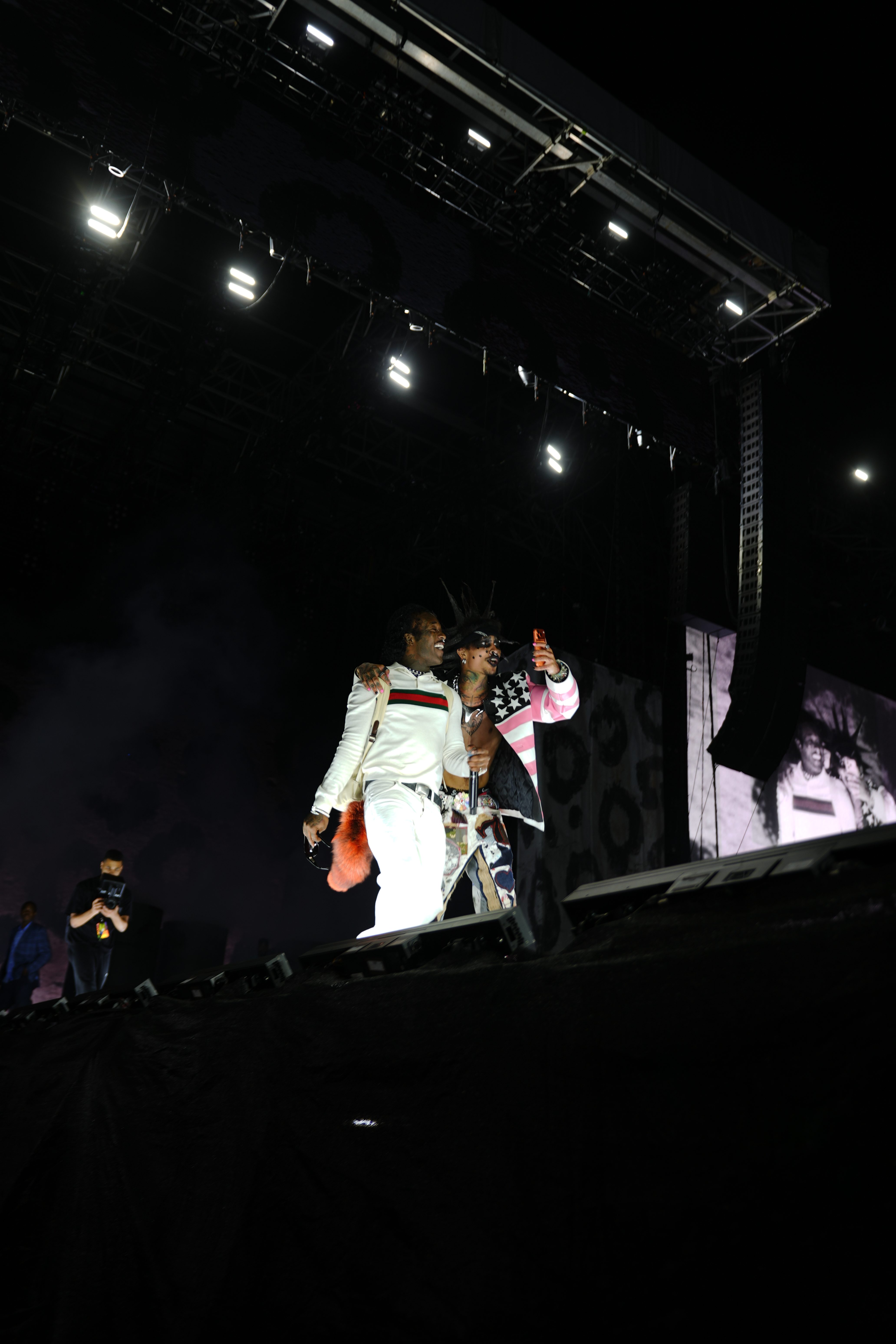 Two performers on a dimly lit stage, one wearing a white and red striped outfit and the other a pink and white outfit with stars, holding microphones and interacting with the audience.