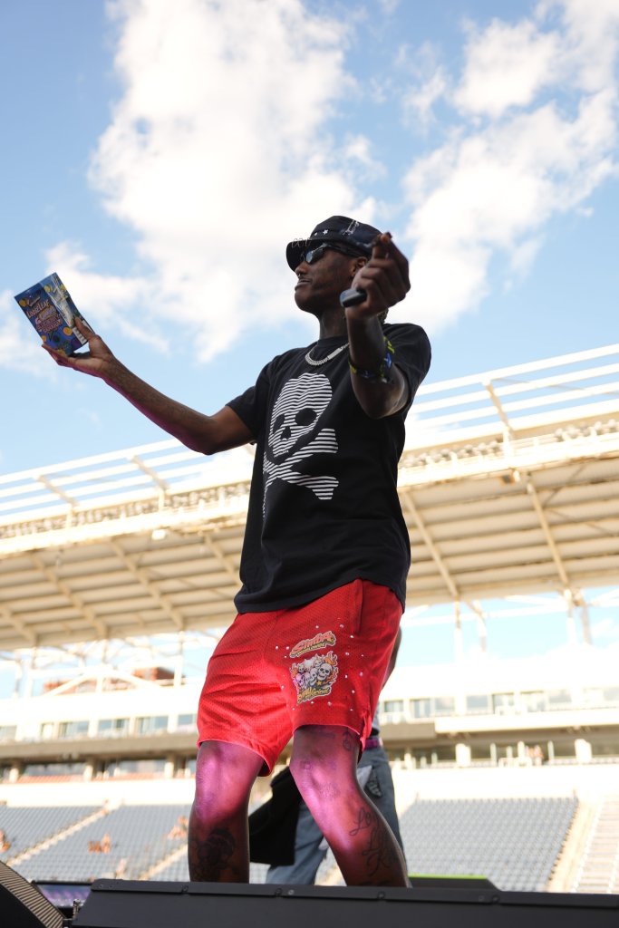 A person wearing a black shirt with a skull graphic, red shorts, and a black hat is holding up a book and looking up at the sky in an outdoor stadium setting.