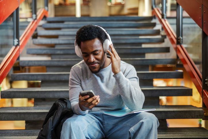 Young black student wearing headphones using smartphone while sitting on stairs