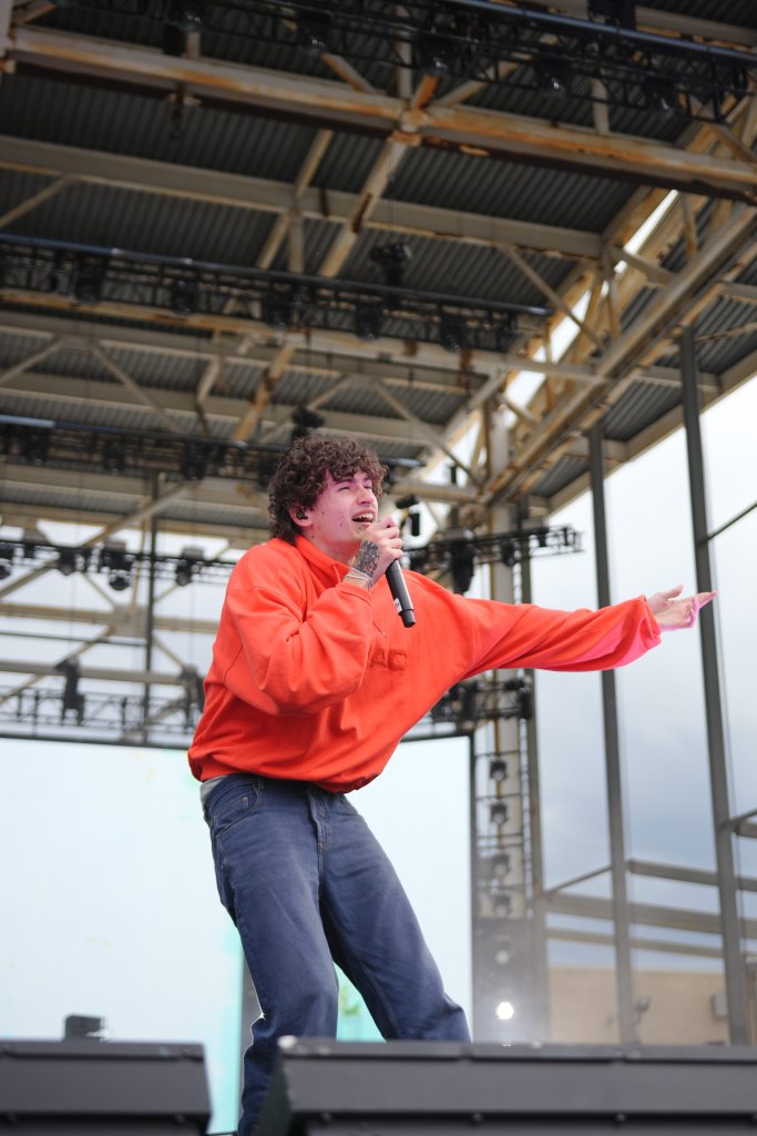 A person wearing a bright red hoodie performing on stage, with a microphone in hand and a metal frame structure visible in the background.