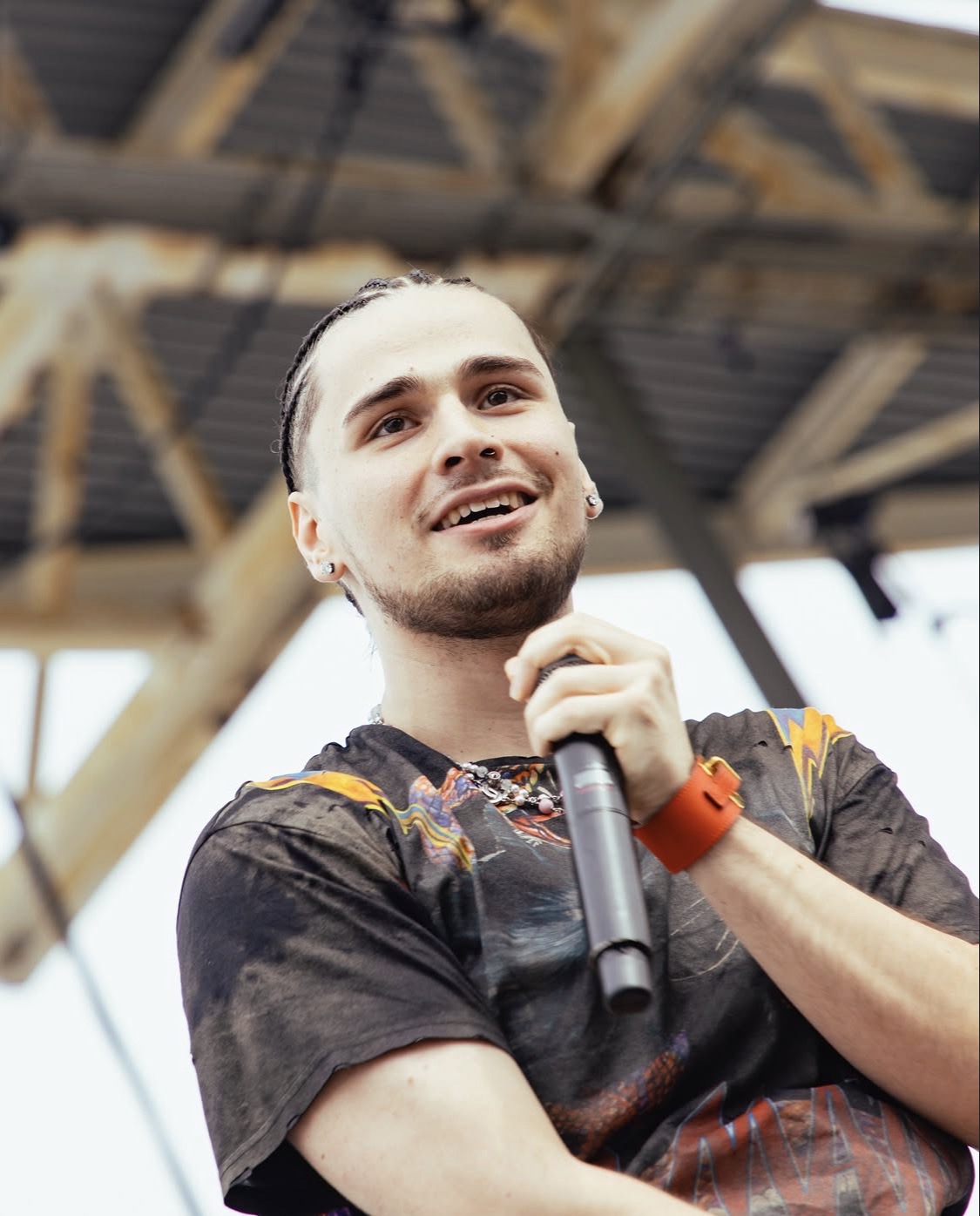 A smiling man holding a microphone on stage, wearing a black t-shirt with a colorful graphic design.