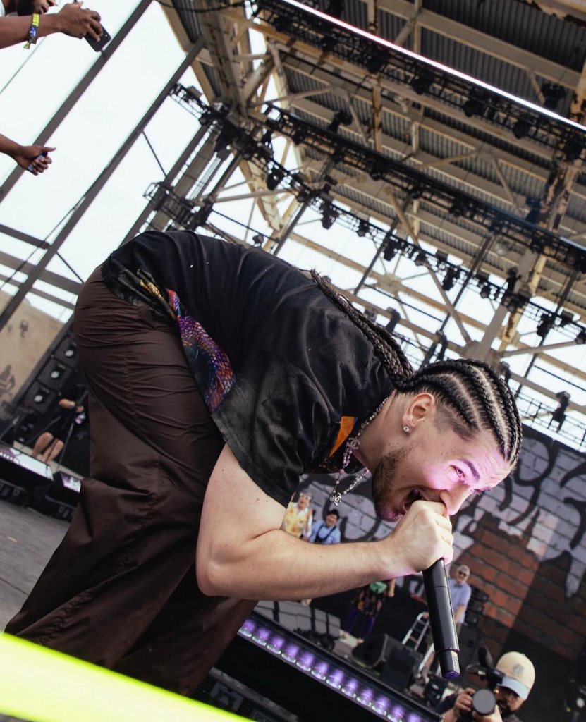 A person in a black outfit performing on stage, with lighting equipment and scaffolding visible in the background.