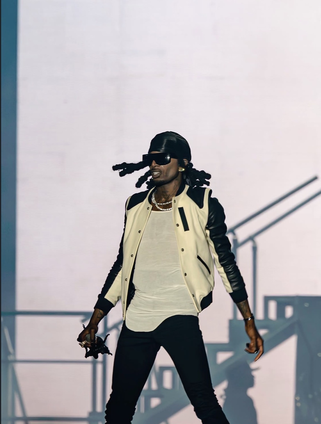 A person wearing a black leather jacket, white shirt, and black pants, with dreadlocks, standing on a stage and holding a microphone.
