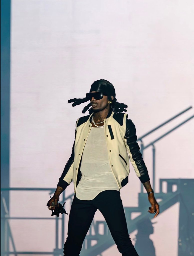 A person wearing a black leather jacket, white shirt, and black pants, with dreadlocks, standing on a stage and holding a microphone.