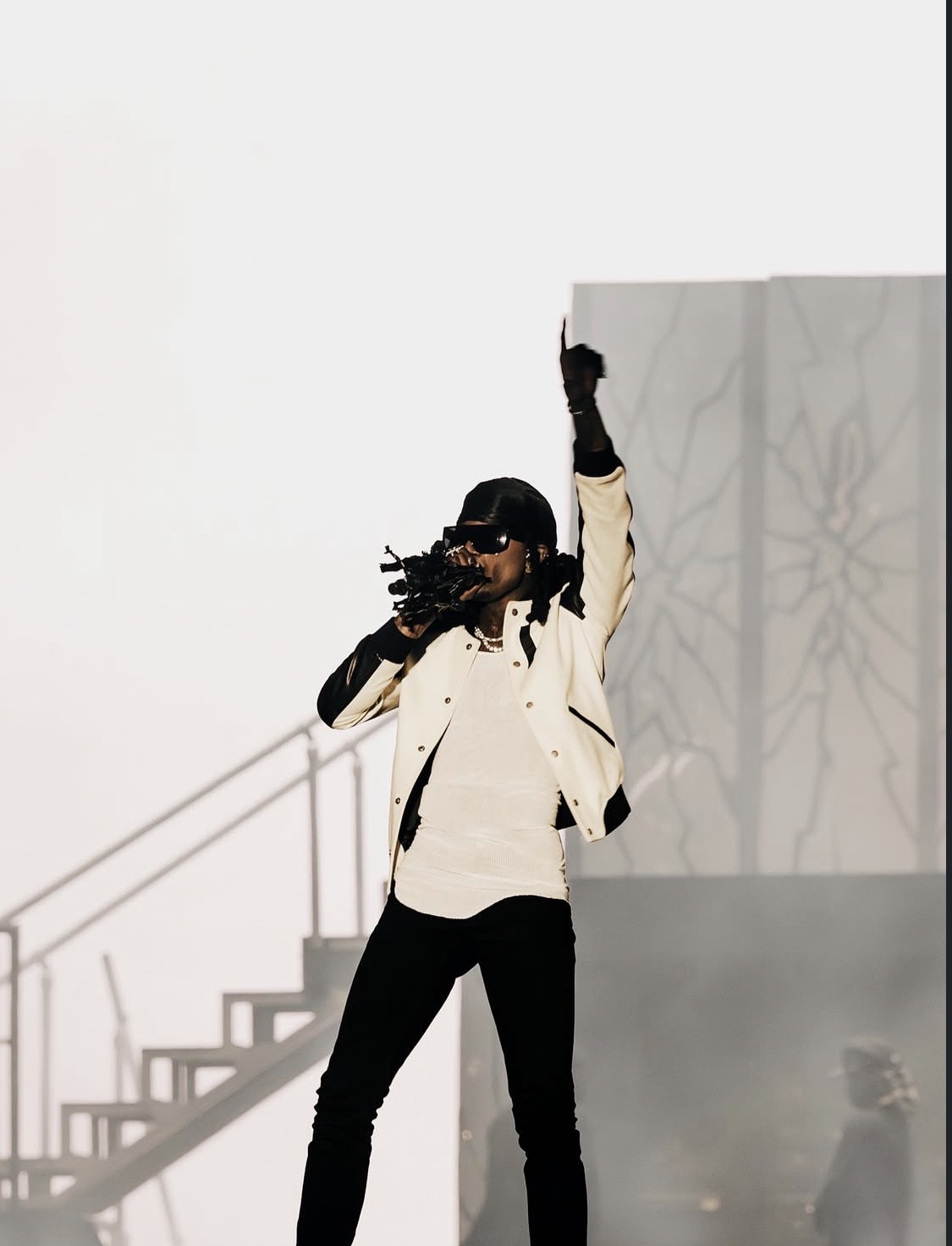 A person wearing a white jacket and black pants stands with their arms raised, holding a camera or other device, in a dimly lit setting with stairs and a white wall in the background.