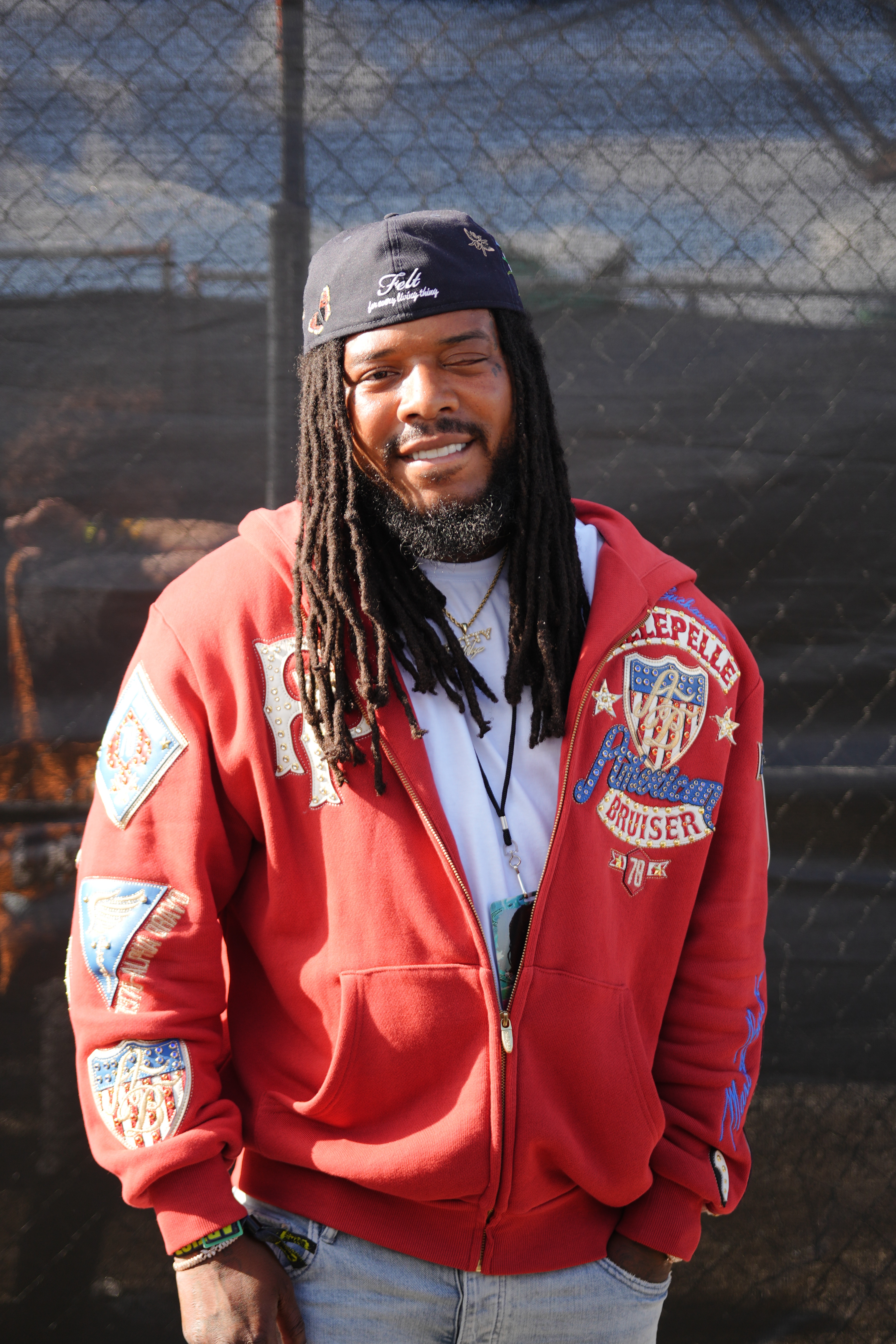 A smiling man with dreadlocks wearing a red jacket with patches and a black hat that says "Felt" stands in front of a chain-link fence.