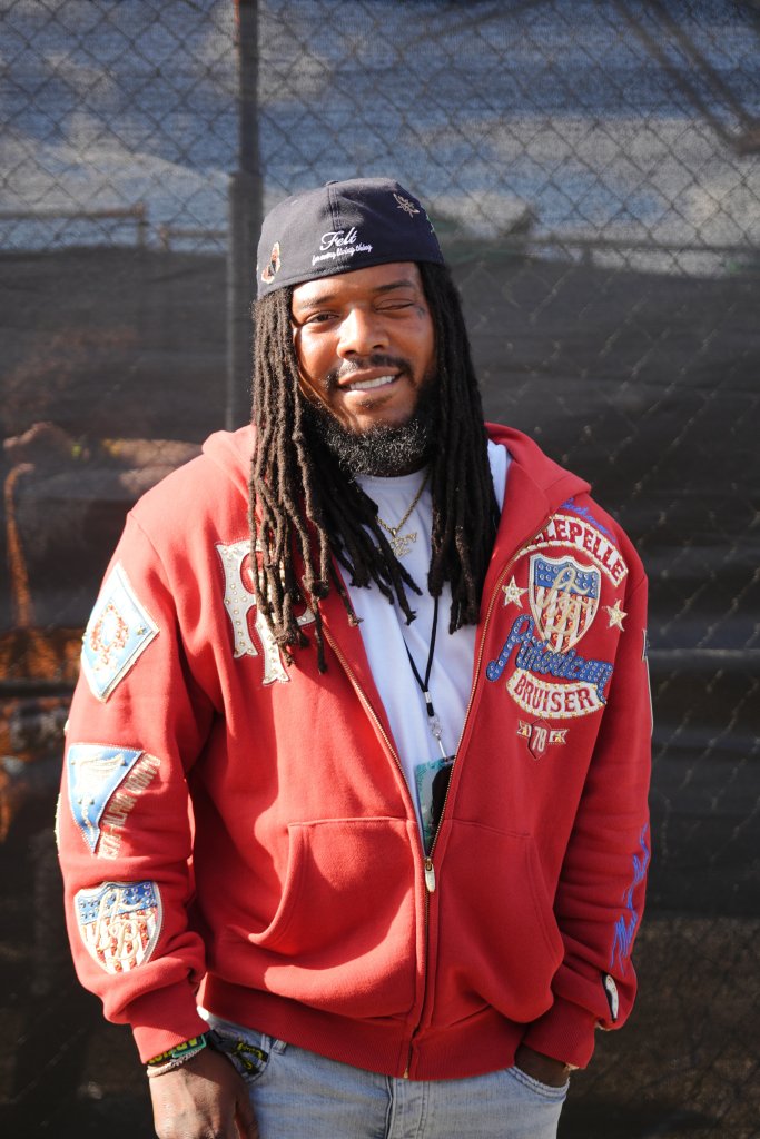 A smiling man with dreadlocks wearing a red jacket with patches and a black hat that says "Felt" stands in front of a chain-link fence.