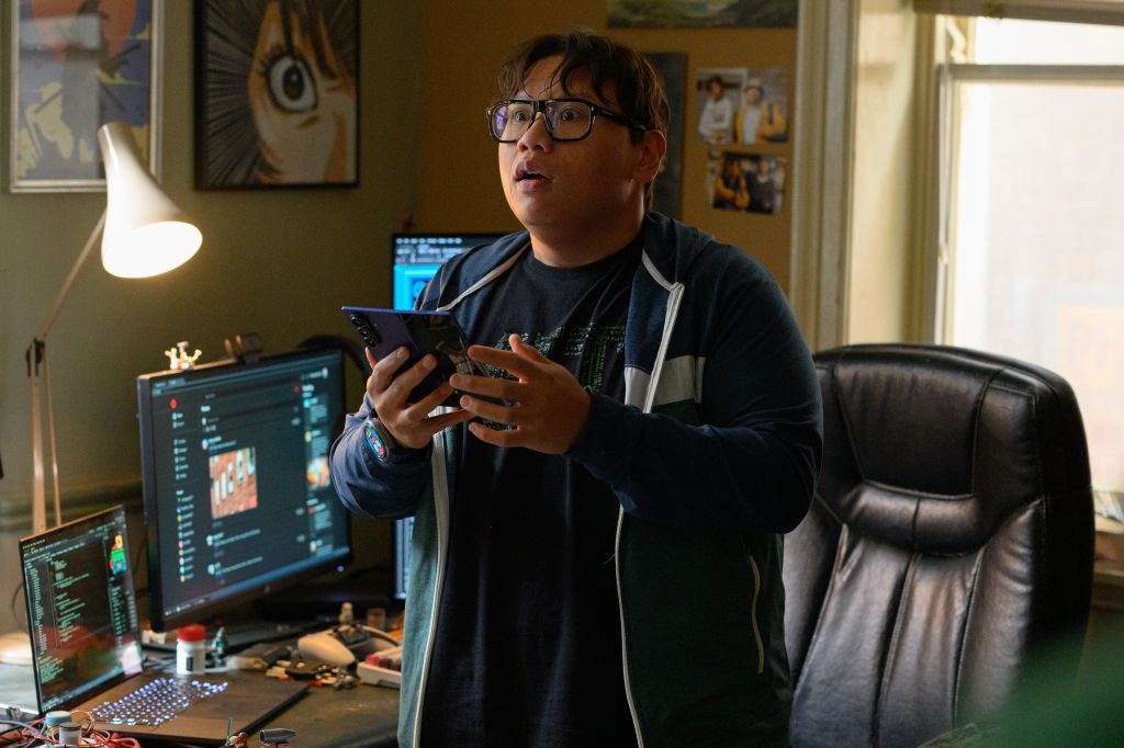 A young person wearing glasses and a black shirt, sitting at a desk with a computer monitor and other electronics, using a smartphone.
