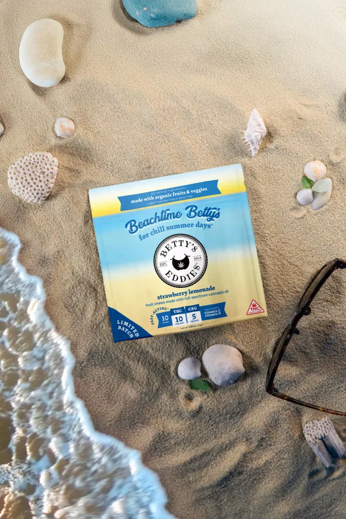 A box of Betty's Eddies strawberry lemonade edibles on a sandy beach with seashells and sunglasses.