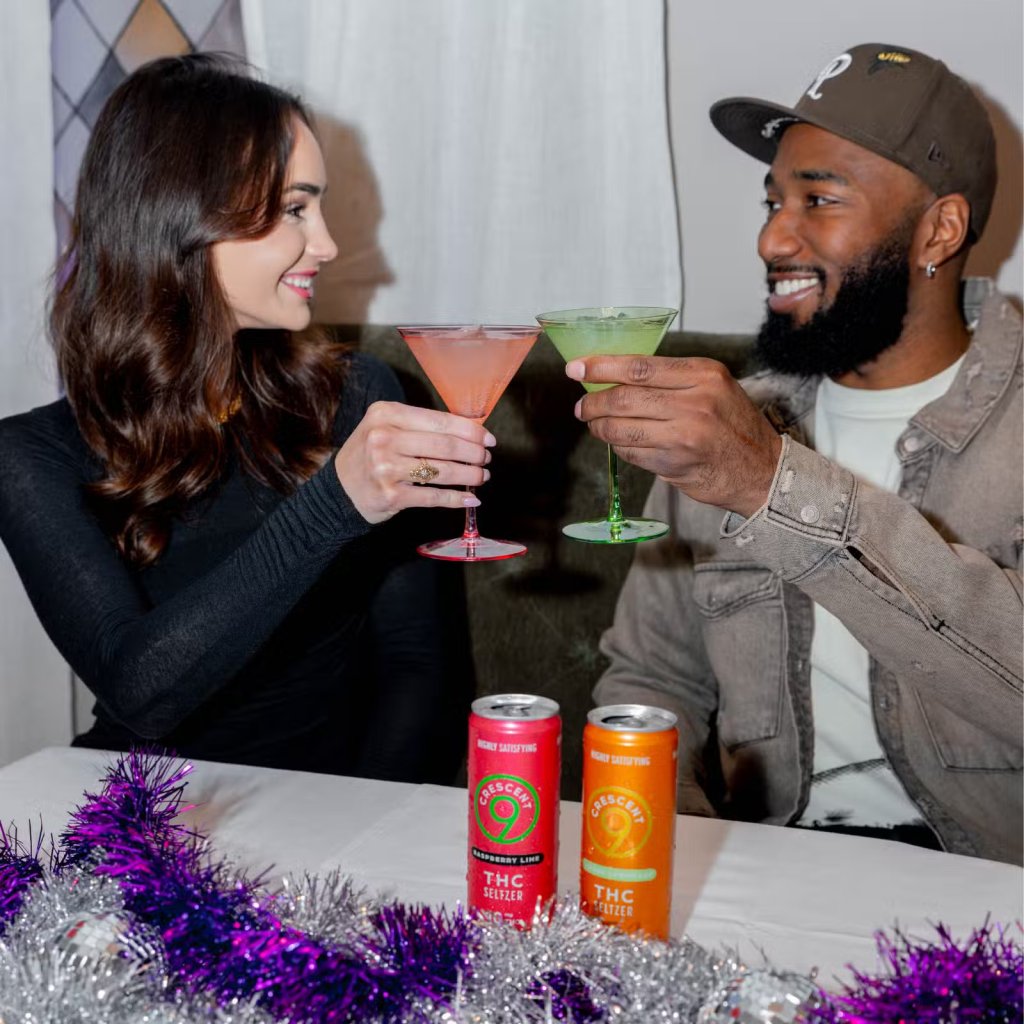 Two people, a woman and a man, holding colorful cocktail glasses and smiling at each other. Cans of THC seltzer are visible on the table.