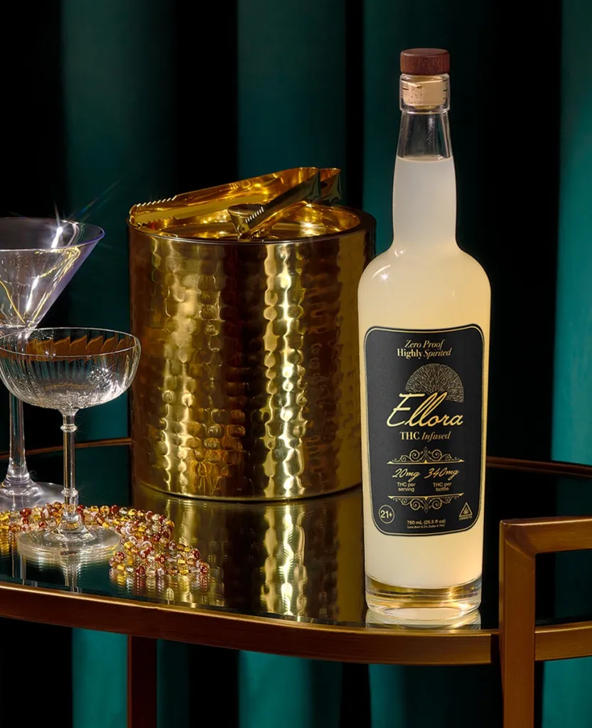A golden, hammered metal ice bucket and a bottle of Ellora THC-infused vodka on a wooden shelf, with a glass of clear liquid in the foreground.