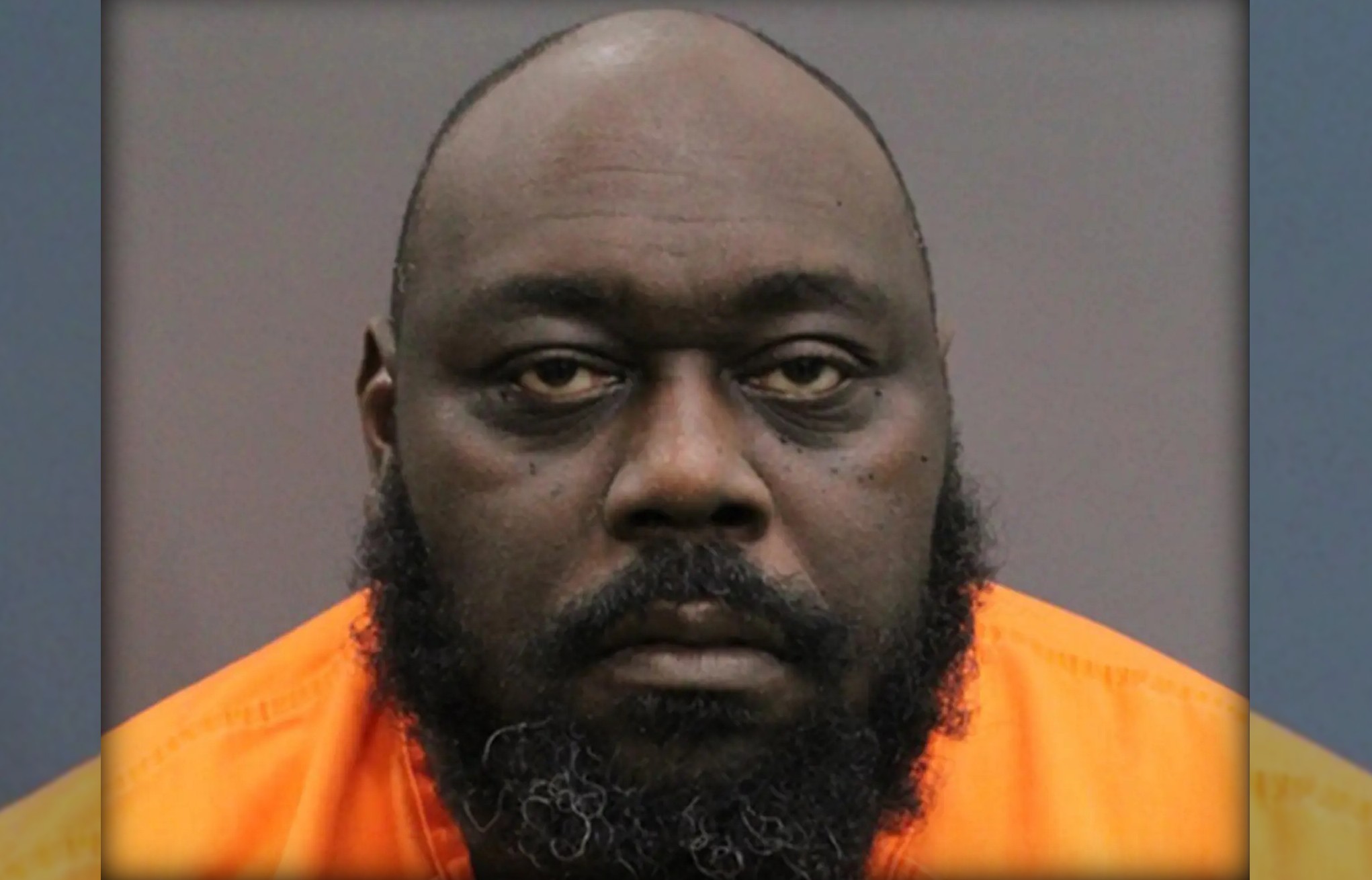 A close-up portrait of a bald, bearded Black man with a serious expression, wearing an orange prison uniform.