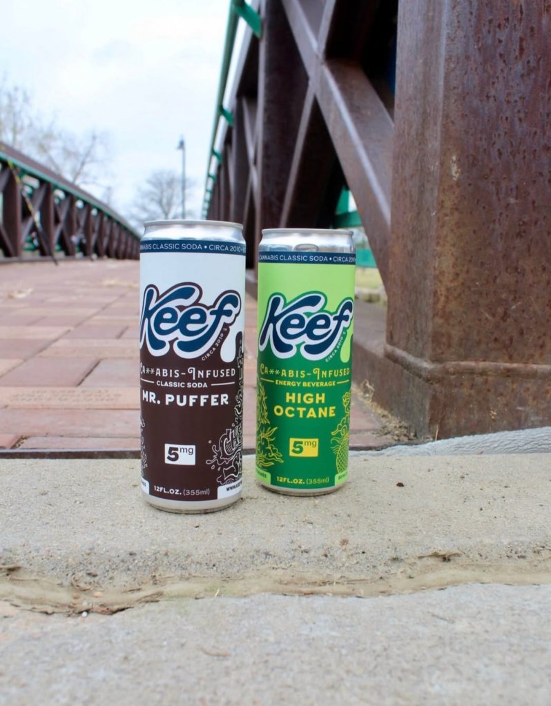 Two cans of Keef cannabis-infused classic soda, one labeled "Mr. Puffer" and the other "High Octane", sitting on a concrete surface in front of a metal structure.
