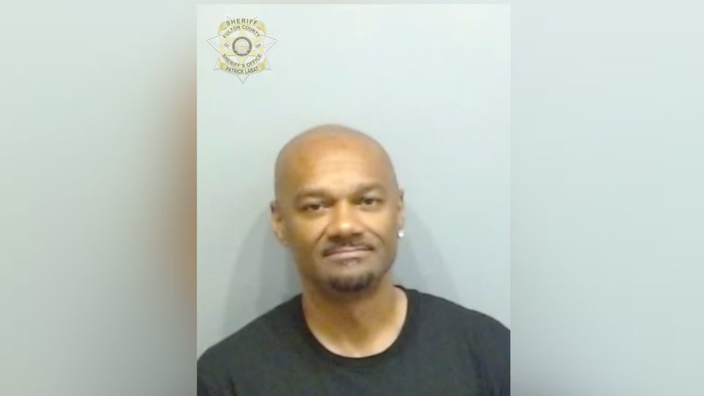Mugshot of a middle-aged bald Black man with a serious expression, wearing a black shirt, against a white background with a sheriff's office badge.