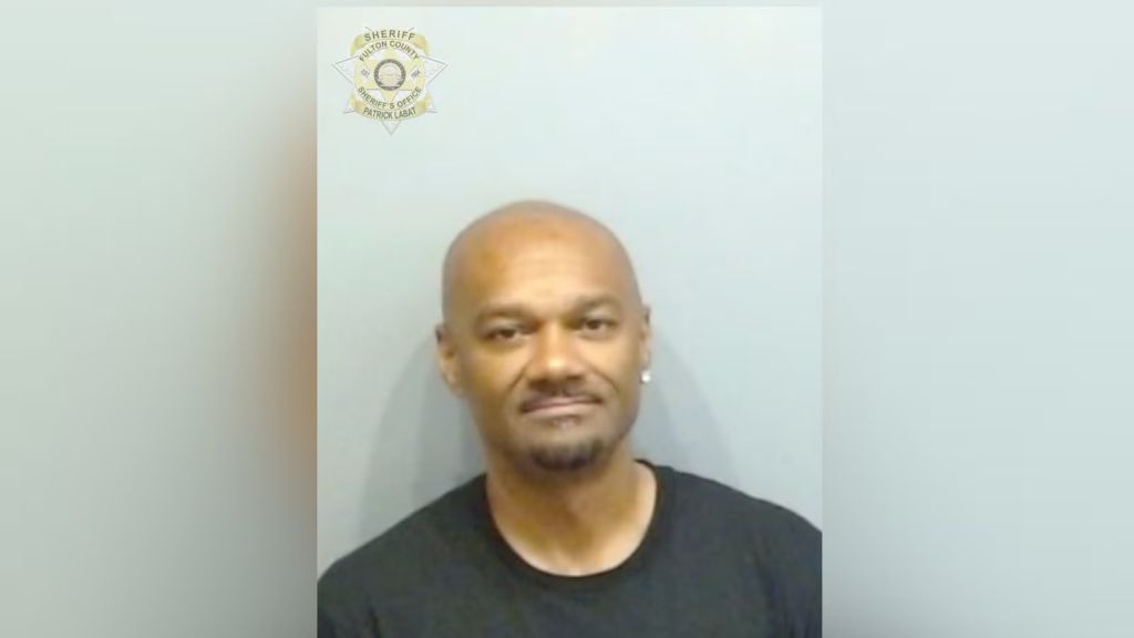 Mugshot of a middle-aged bald Black man with a serious expression, wearing a black shirt, against a white background with a sheriff's office badge.