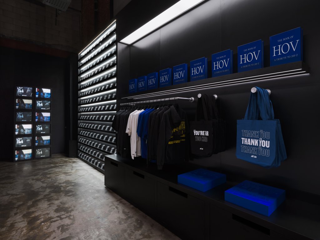 Shelves of books titled "The Book of HOV" in a dark retail space, with clothing and tote bags on display.