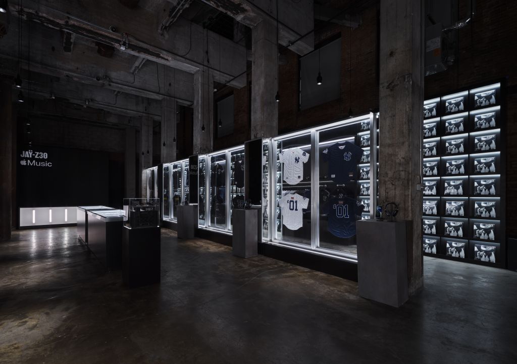 A dark, industrial-style room with shelves displaying various sports jerseys and memorabilia behind glass panels. The room is illuminated by bright lighting, creating a dramatic, museum-like atmosphere.
