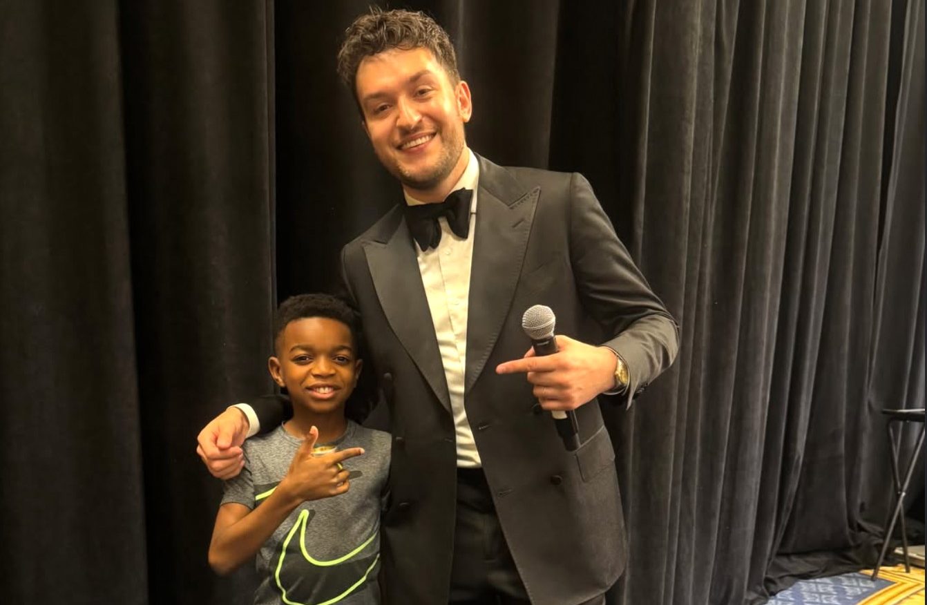 A man in a suit holding a microphone, standing next to a smiling young boy in a gray t-shirt and black shorts.