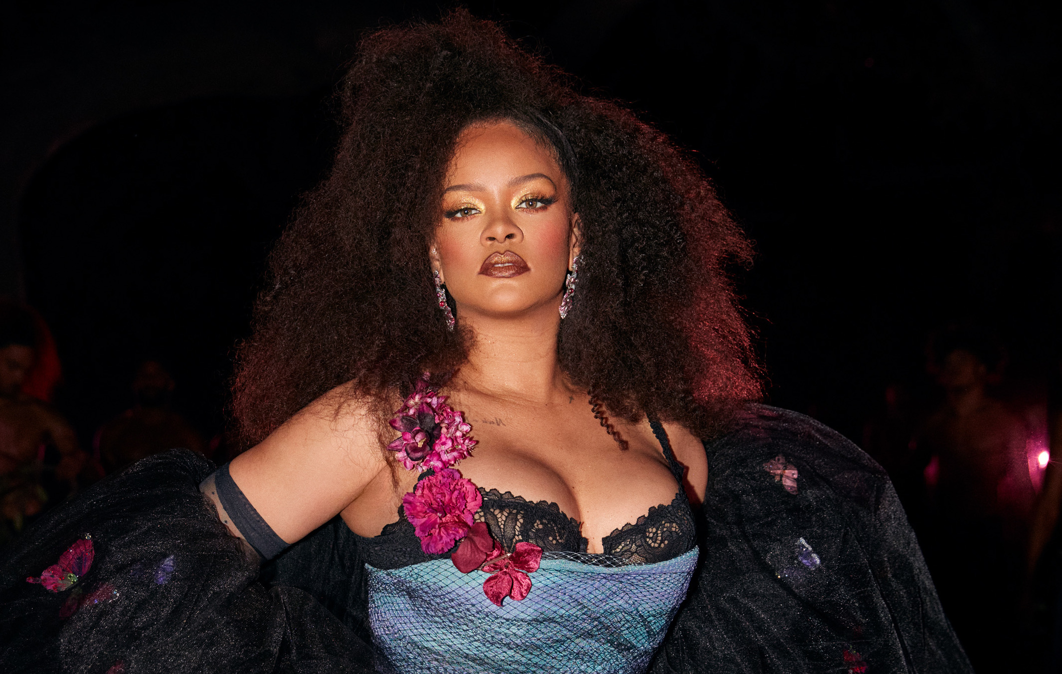 A woman with curly dark hair wearing a black lace bra and a black and blue patterned jacket, posing with a confident expression.