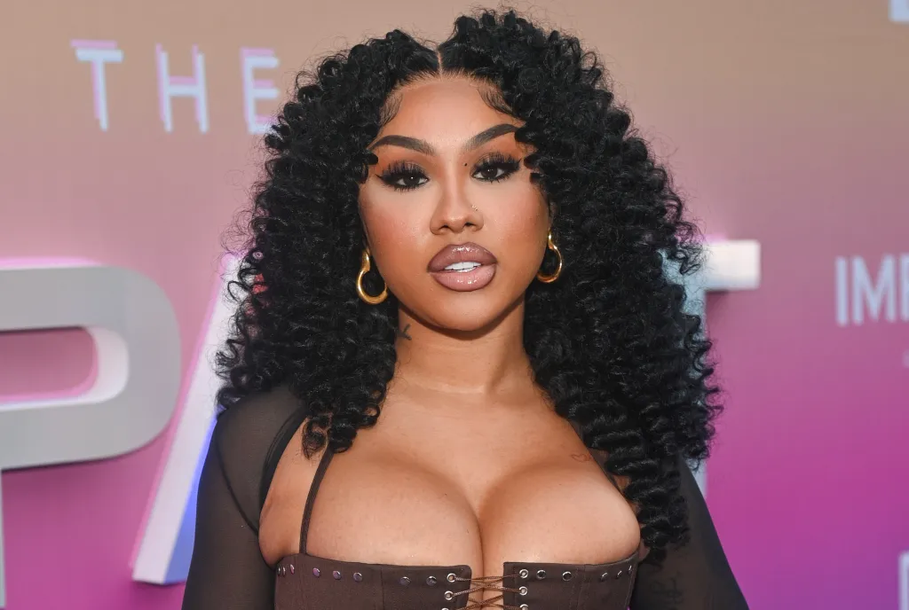 A woman with curly black hair wearing a black outfit and large hoop earrings poses in front of a pink and white backdrop with the text "THE IMPACT ATLANTA".