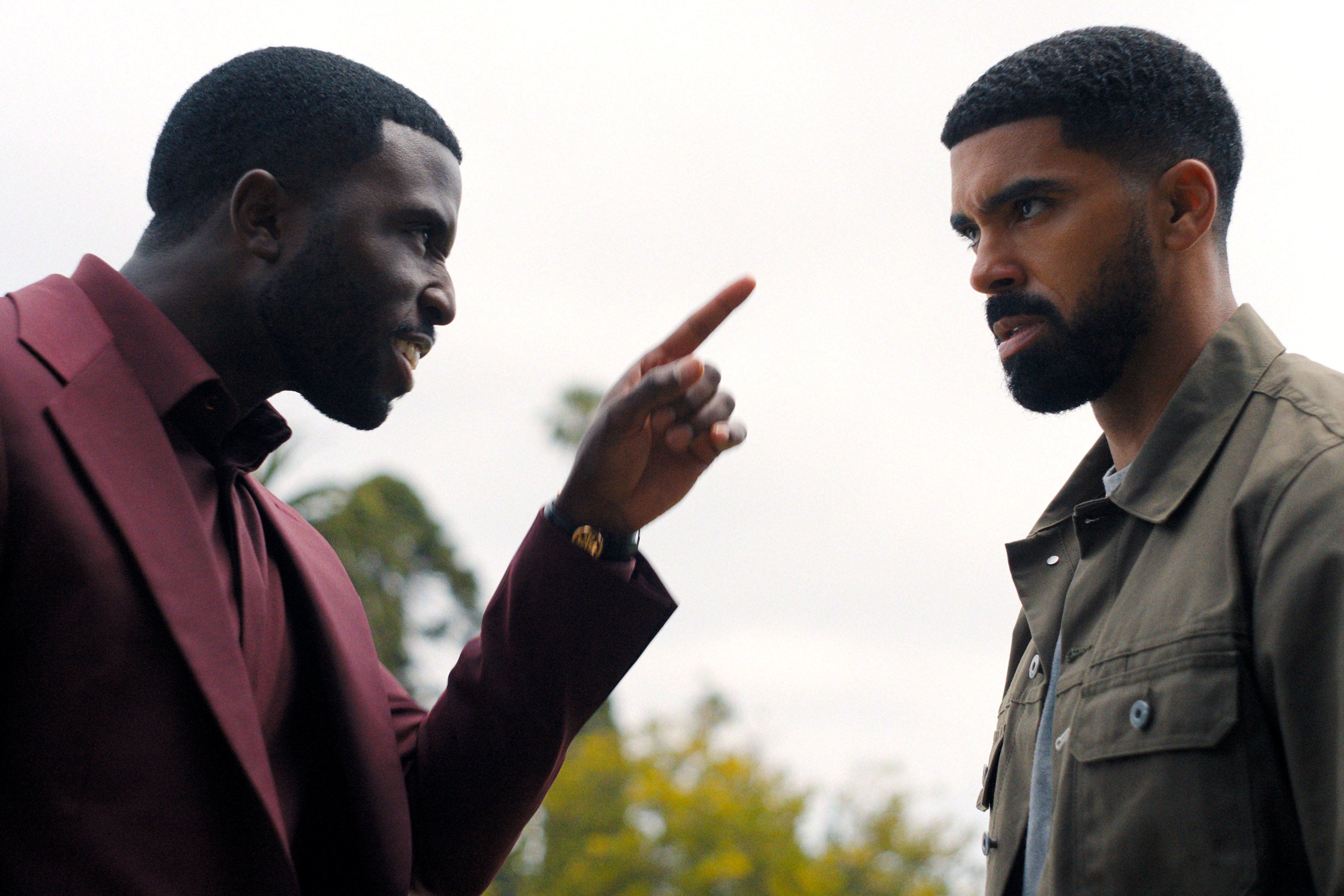 "Two men outdoors in a confrontation, one angrily pointing his finger at the other, who looks serious and tense; trees and sky are visible in the background."