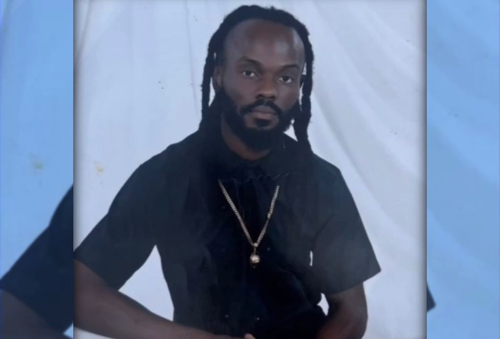 A man with dreadlocks wearing a black shirt and necklace, looking directly at the camera.