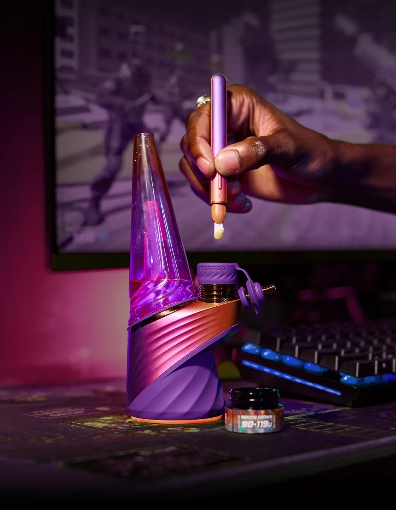 A person's hand holding a purple and pink vaping device, with a keyboard and other electronic devices in the background.