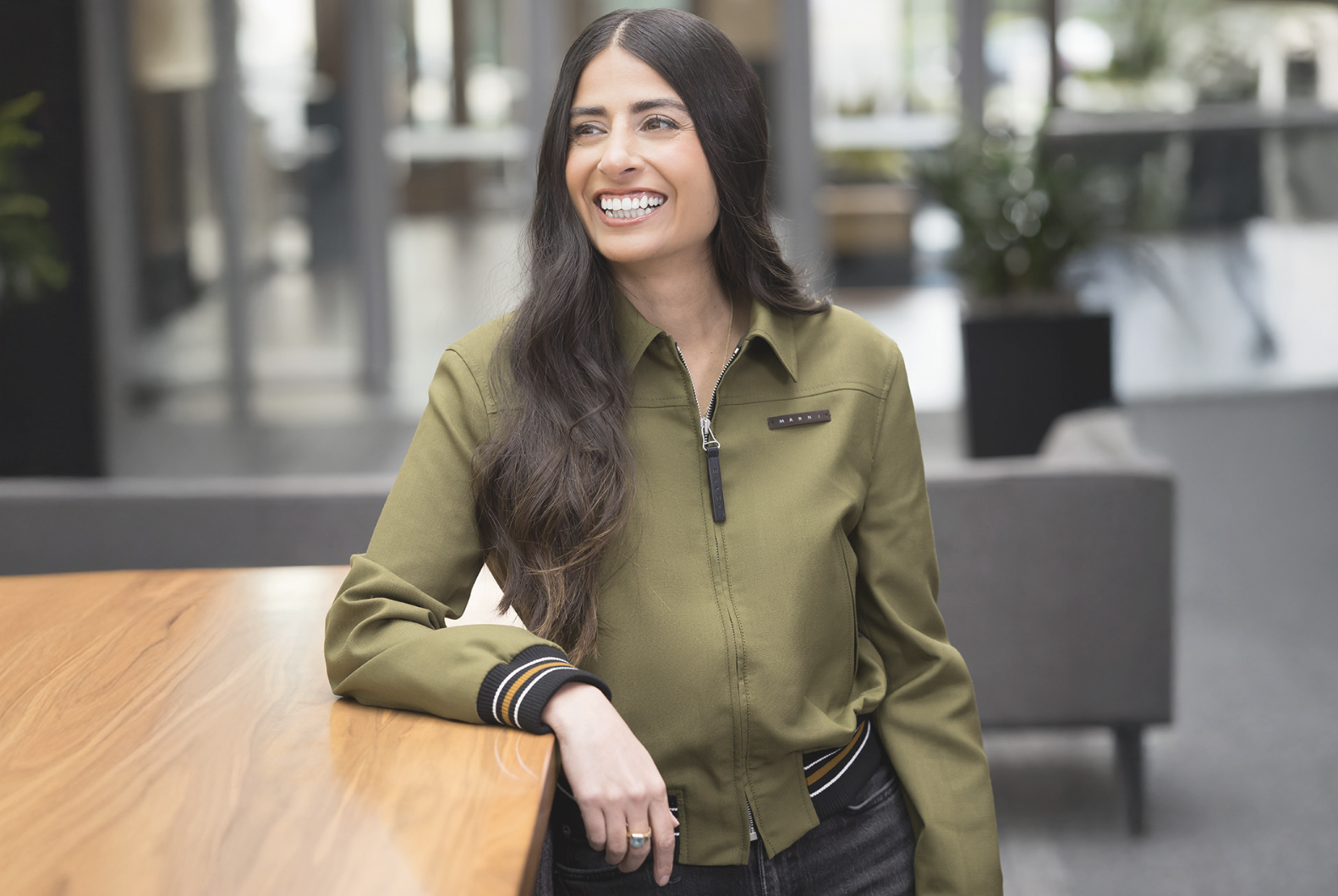 A smiling woman with long dark hair wearing a green jacket stands in front of a glass building.