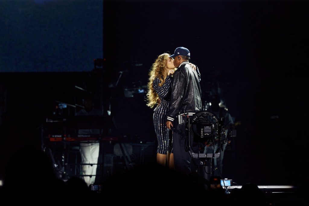Two people embracing on a dimly lit stage, with a camera and other production equipment visible in the background.