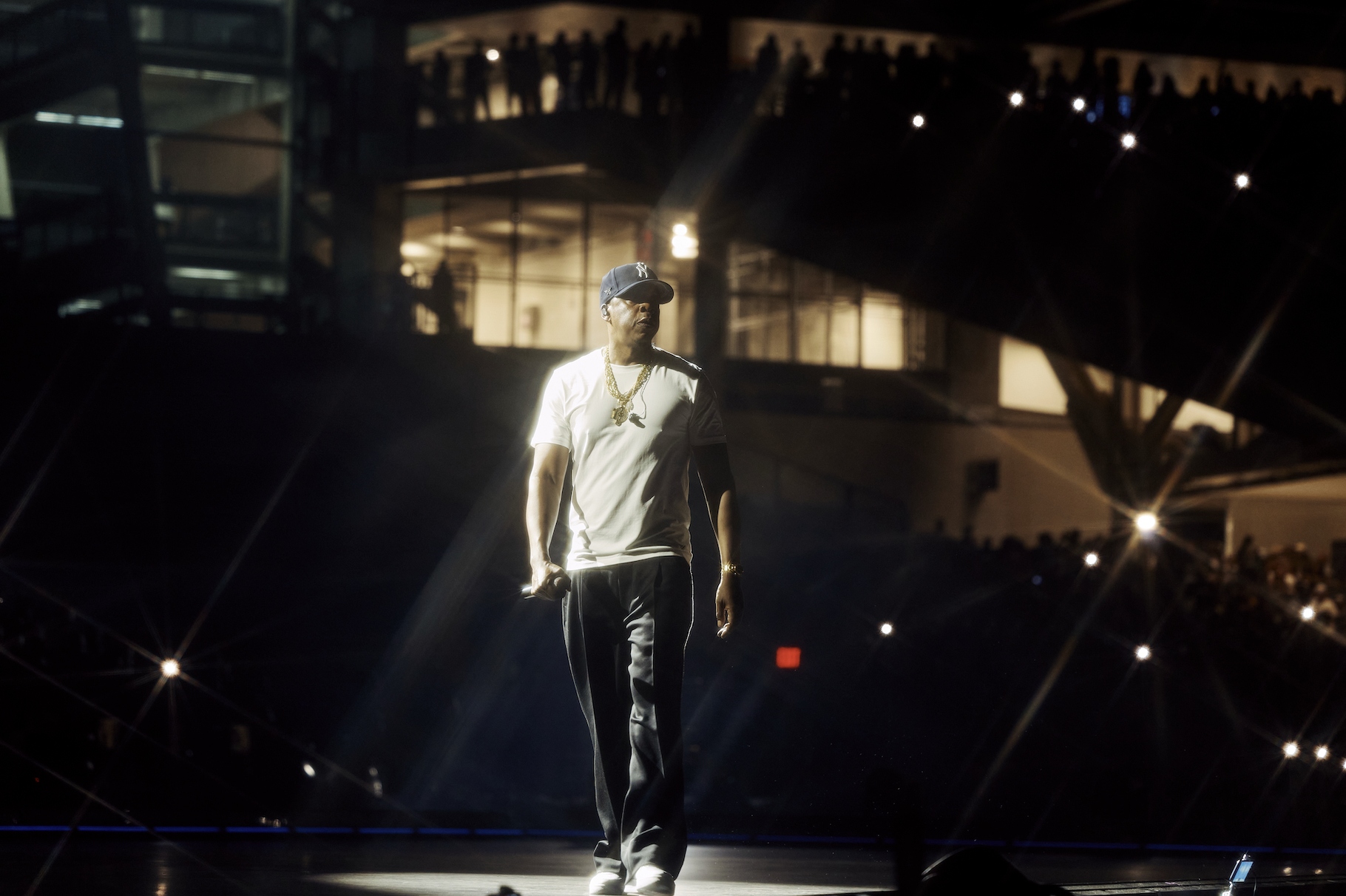 A person wearing a white shirt, baseball cap, and necklace standing on a stage in a dimly lit venue.